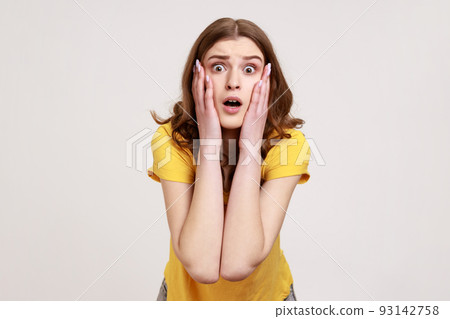 Astonished teenager girl with brown in casual yellow T-shirt standing with mouth open in surprise, has shocked expression, keeps palms on cheeks. Indoor studio shot isolated on gray background. Astonished teenager girl with brown in casual yellow T-shirt standing with mouth open in surprise, has shocked expression, keeps palms on cheeks. Indoor studio shot isolated on gray background. 93142758