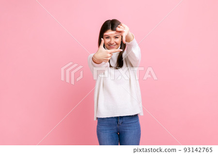 Smiling woman looking through finger frame, imitating to take photo, focusing interesting moment, wearing white casual style sweater. Indoor studio shot isolated on pink background. 93142765