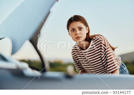 A sad woman looks in disbelief at her broken down car on the road while traveling and does not know what to do 93144177