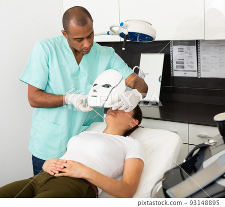 Beautician preparing female client for face phototherapy Beautician preparing female client for face phototherapy 93148895