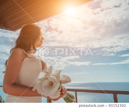 Gorgeous brunette bride posing with a bouquet on hotel terrace, looking at sea. Luxury destination, wedding makeup and hairstyle. Morning of the bride. Puffy lips and expressive eyes. Wedding concept 93152302