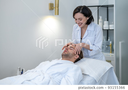 Dark-haired professional doing face massage to a male customer Dark-haired professional doing face massage to a male customer 93155366