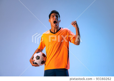 Portrait of young emotive man, motivated football player in uniform with ball posing isolated over blue studio background in neon light Portrait of young emotive man, motivated football player in uniform with ball posing isolated over blue studio background in neon light 93160262