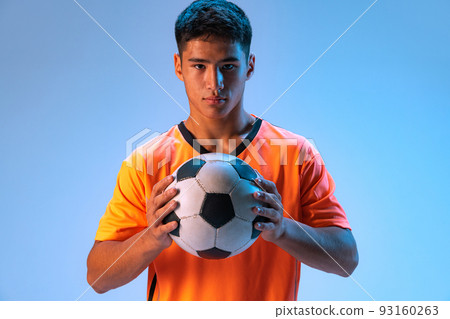 Portrait of young man, professional football player in uniform posing with ball, looking at camera isolated over blue background in neon light 93160263