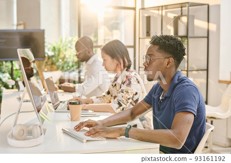African young programmer in eyeglasses concentrating on his work on computer sitting at table with his colleagues at office 93161192