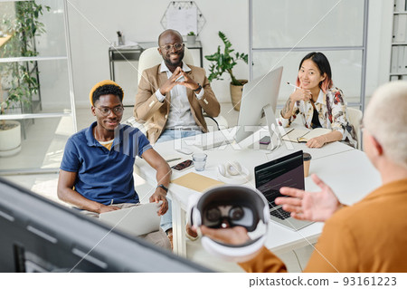 Multiethnic group of colleagues sitting at table at meeting and listening to speaker telling about new app of virtual reality glasses 93161223