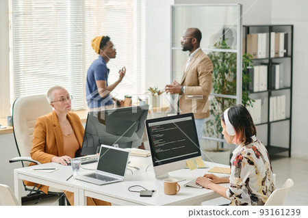 Two colleagues working with codes on computers at table with men discussing something in background 93161239
