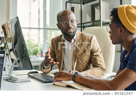African manager discussing new computer project with young programmer at table at office 93161247