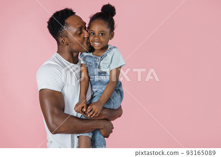 Vertical portrait of happy afro daddy embracing with his cute baby daughter, pink studio background. Vertical portrait of happy afro daddy embracing with his cute baby daughter, pink studio background. 93165089