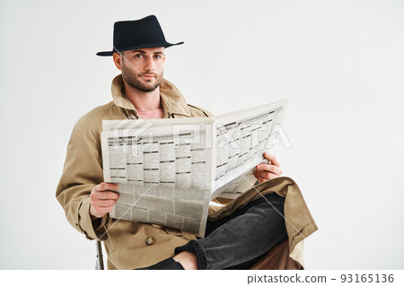Young handsome man in a trench coat and hat reading newspaper sitting on chair over white background Young handsome man in a trench coat and hat reading newspaper sitting on chair over white background 93165136