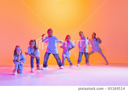 Dance group of happy, active little girls in t-shirts and jeans in action isolated on orange background in neon. Concept of music, fashion, art, childhood Dance group of happy, active little girls in t-shirts and jeans in action isolated on orange background in neon. Concept of music, fashion, art, childhood 93169514