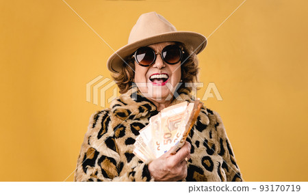 Happy senior woman holding and showing money banknotes while standing over isolated background. 93170719