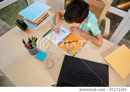 Top view. School boy solves maths and geometry examples, sitting at a table with school supplies, measuring rulers, stacked textbooks, black alarm clock and laptop, broadcasting an online video lesson 93171265
