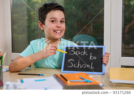 Cute schoolboy points with pencil on a chalkboard with inscription BACK TO SCHOOL, sitting at table with textbooks and school supplies, cutely smiles looking at the camera. New academic year semester Cute schoolboy points with pencil on a chalkboard with inscription BACK TO SCHOOL, sitting at table with textbooks and school supplies, cutely smiles looking at the camera. New academic year semester 93171266