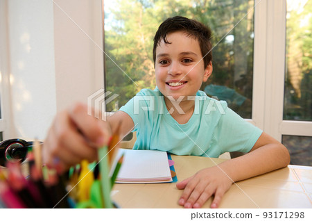 Handsome school boy of 10 years old taking out a pen from pencil holder while sitting at desk and doing his homework, smiles a cheerful toothy smile, expressing sincere positive emotionsBack to school 93171298