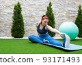 young woman with red hair, practising leg stretching exercises on a mat in the garden of her house. Sporty girl training. concept of health and well-being. 93171493
