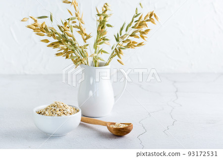 Dry oatmeal in a bowl and in a wooden spoon and a jug with ears on a light table. Home healthy food Dry oatmeal in a bowl and in a wooden spoon and a jug with ears on a light table. Home healthy food 93172531