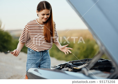 A woman traveler stands by her old car with the hood open and repairs the car engine with a wrench, unscrewing car parts 93173208