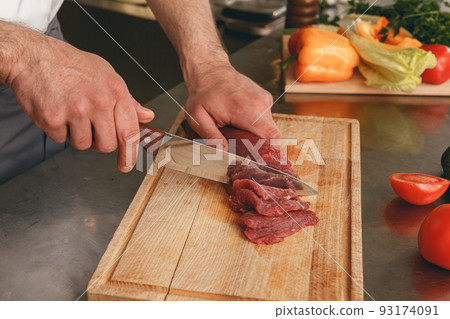Close up of chef cutting fresh raw meat on wooden board in modern kitchen of restaurant Close up of chef cutting fresh raw meat on wooden board in modern kitchen of restaurant 93174091