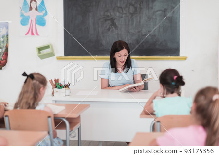 A teacher reads a book to elementary school students who listen carefully while sitting in a modern classroom A teacher reads a book to elementary school students who listen carefully while sitting in a modern classroom 93175356