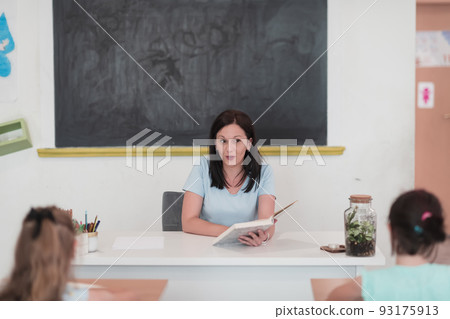 A teacher reads a book to elementary school students who listen carefully while sitting in a modern classroom A teacher reads a book to elementary school students who listen carefully while sitting in a modern classroom 93175913
