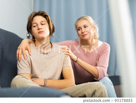 Upset and offended teenager sitting on sofa while mother soothing him Upset and offended teenager sitting on sofa while mother soothing him 93178881