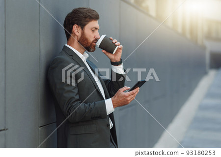 Focused european businessman in suit with phone drinking coffee near office building during break Focused european businessman in suit with phone drinking coffee near office building during break 93180253