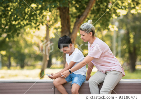Mother trying to take a mobile phone away from her 10 years old son, sitting on the bench outdoor in public park in summer, selective focus. 93180473