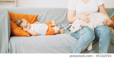 A girl sitting on a sofa strokes a puppy on her lap next to a little girl A girl sitting on a sofa strokes a puppy on her lap next to a little girl 93182050