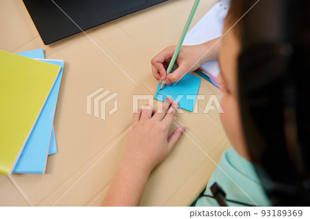 Overhead view of a schoolchild during a video conference with the teacher in virtual class, with selective focus on his hands holding a pencil and making notes during an online lesson on the laptop 93189369