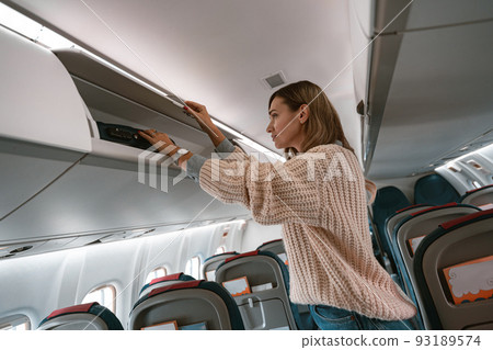 Woman traveler putting luggage into overhead locker on airplane during boarding Woman traveler putting luggage into overhead locker on airplane during boarding 93189574