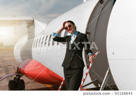 Woman flight attendant in sunglasses standing on airplane stairs at airport and looking camera 93189645