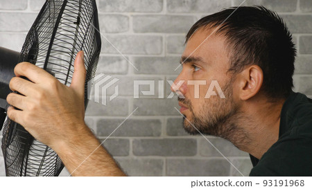 Close-up of a bearded man looking at a fan. The man brings his face to the fan. In the background is a stone wall of light color. Side view. The concept of hot weather. 93191968