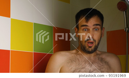 A man turns on the cold water in the shower and squirms his face from the cold. Caucasian man takes a shower on a hot summer day. There are colorful tiles in the background. 93191969