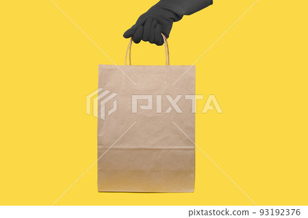 Close up female holds in hand brown clear empty blank craft paper bag for takeaway isolated on yellow background. Packaging template mock up. Delivery service concept. Copy space. Advertising area 93192376