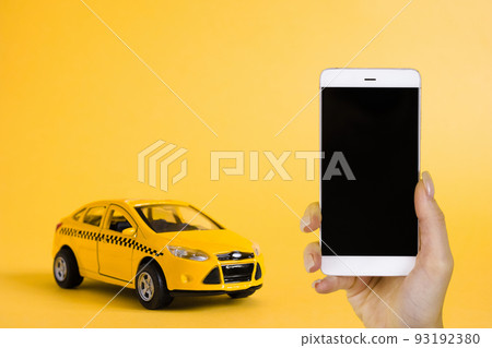 Urban taxi mobile online application concept. Toy yellow taxi car model. Hand holding smart phone with taxi service app on display. Mock up with copy space. 93192380