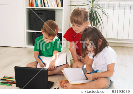 Children sit on the floor with pens and notebooks and use a laptop 93193089