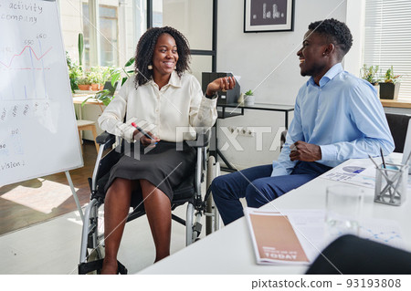 African businesswoman sitting on wheelchair near the flipchart and discussing business strategy with her partner at office African businesswoman sitting on wheelchair near the flipchart and discussing business strategy with her partner at office 93193808