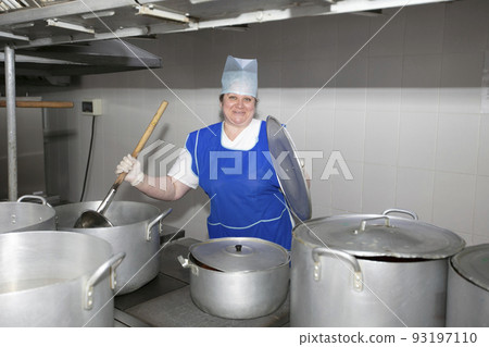 A woman cook in an industrial kitchen with a large ladle among metal pans. Chef at the hospital A woman cook in an industrial kitchen with a large ladle among metal pans. Chef at the hospital 93197110