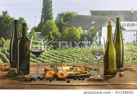 Bottles and glasses of red and white wine with delicious appetizers, olives, cheese and honey, fruits and berries lying on table. Countryside vineyard view 93198695