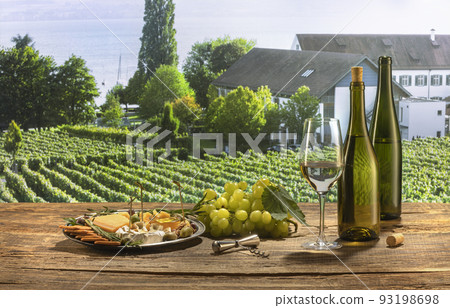 Two bottles and glass of delicious white wine standing on wooden table. Yummy appetizers with cheese, honey, grapes and crackers. Summer vineyard landscape view 93198698
