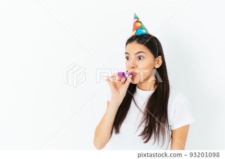 Portrait of funny young woman wearing birthday cap blowing kazoo 93201098