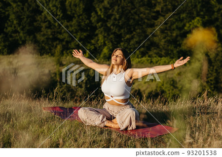 Young Woman Sunbathing and Meditating on the Mat Outdoors at Sunset with Beautiful Landscape on the Background Young Woman Sunbathing and Meditating on the Mat Outdoors at Sunset with Beautiful Landscape on the Background 93201538