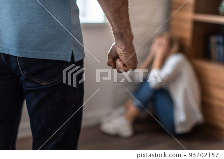 Scared crying mature caucasian woman covers face sits on floor and afraid of aggressive man with fist Scared crying mature caucasian woman covers face sits on floor and afraid of aggressive man with fist 93202157