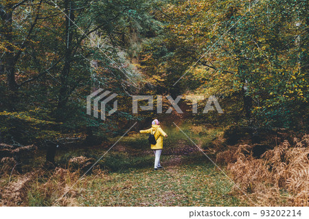 Back view woman with raised arms in bright yellow raincoat enjoying life and freedom in autumn forest in nature. Feeling harmony, reunion with nature in fall. Relaxing, personal fulfillment. 93202214