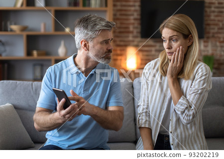 Unhappy adult caucasian man with beard shows phone to shocked lady, woman suffers from gadget addiction 93202219