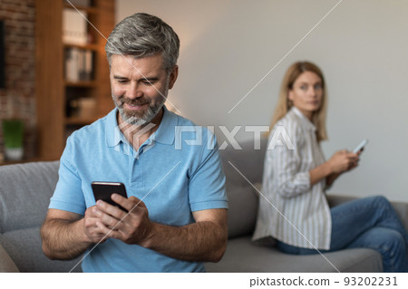 Happy adult caucasian husband with beard plays on smartphone, suffers from gadget addiction, ignores wife 93202231