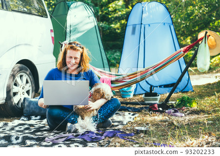 Adult woman use laptop computer sitting with her cockapoo puppy near camper van recreational vehicle with background of tents and nature park. Concept of travel and remote worker people. 93202233