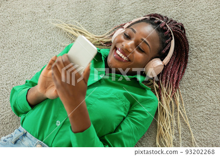 Happy african american lady in headphones lying on floor and using smartphone, listening to music and chatting online Happy african american lady in headphones lying on floor and using smartphone, listening to music and chatting online 93202268