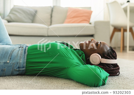 Young relaxed black lady in wireless headphones listening to music or audio book, lying on floor at home, side view 93202303